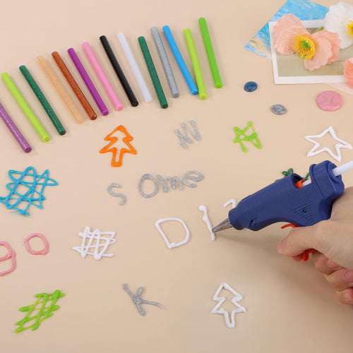 Hand holding a blue hot glue gun creating white glue art on a beige surface, surrounded by colorful mini glue sticks and various glue shapes.