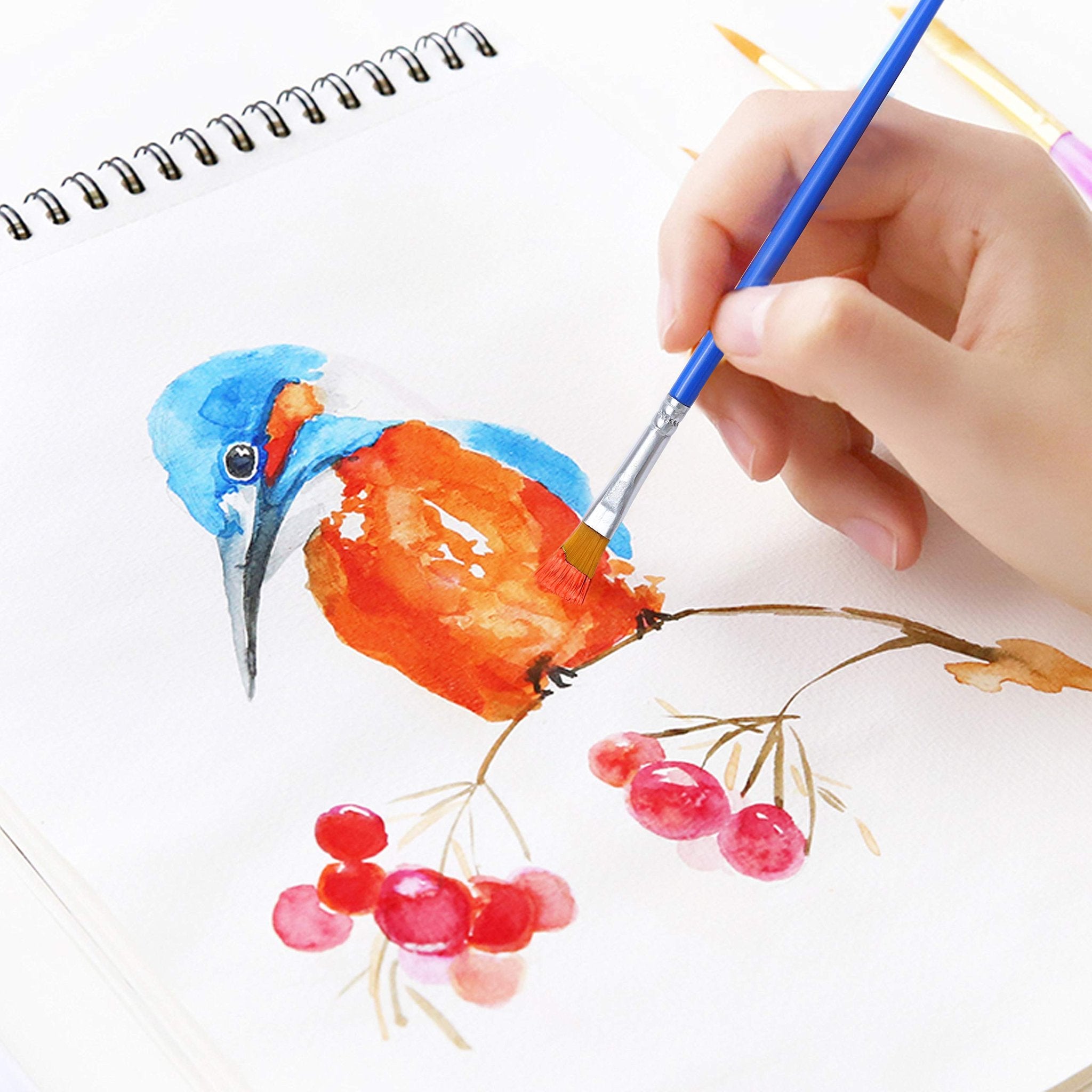 Hand holding a blue paintbrush adding detail to a watercolor painting of a bird on a branch with berries.