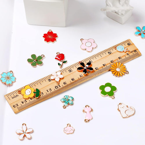 Assortment of colorful enamel charms including flowers, a cat, a cloud, and a lollipop, laid out with a wooden ruler for scale.