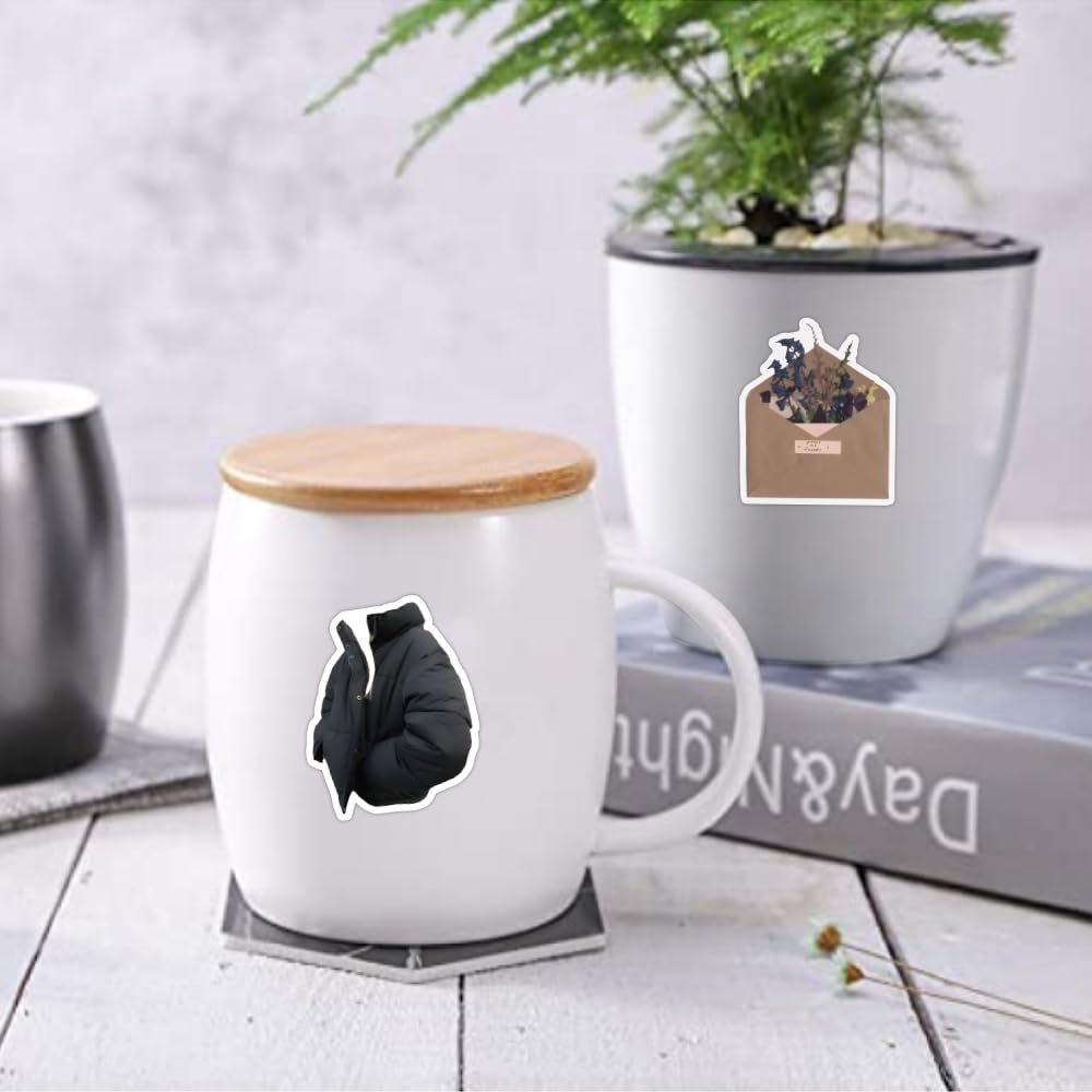 Two aesthetic stickers applied to a white mug and a plant pot: a puffer jacket sticker on the mug and an envelope with dried flowers sticker on the pot.