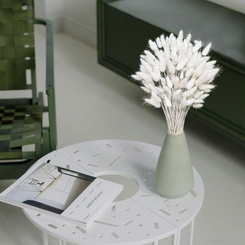 Bouquet of white dried pampas grass in a sage green vase on a white table with a magazine