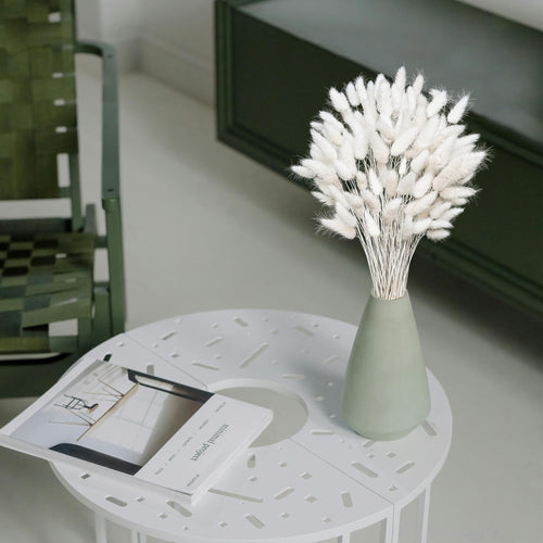 Bouquet of white dried pampas grass in a sage green vase on a white table with a magazine