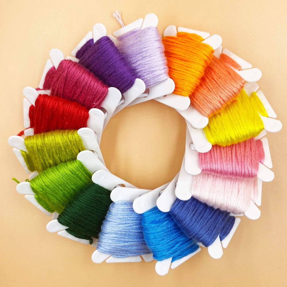 Circular arrangement of white plastic bobbins holding colorful embroidery floss on a light peach background.