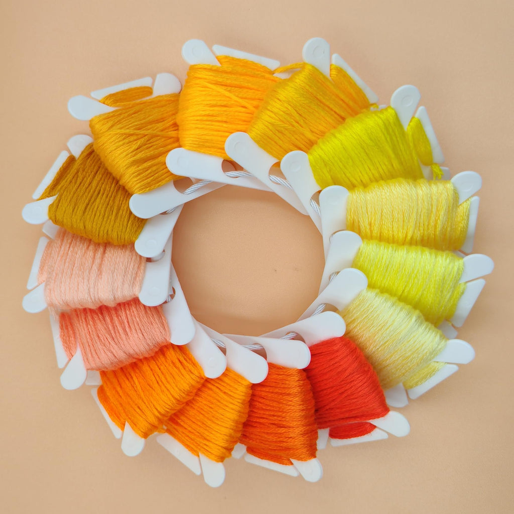 Embroidery floss bobbins arranged in a circle on a peach background, showing shades of yellow, orange, and pink.