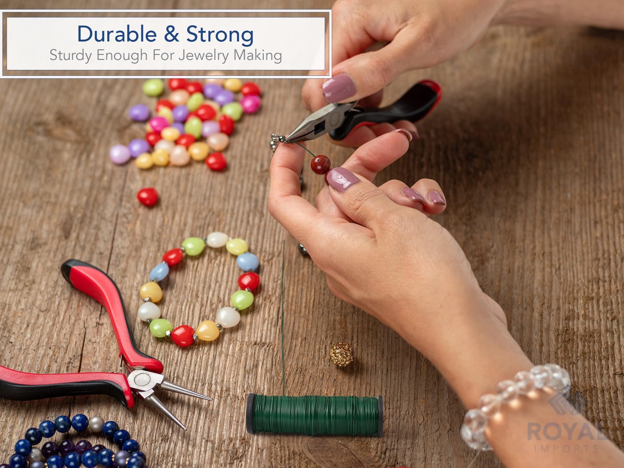 Hands using pliers to thread beads onto green floral wire spool on wooden surface with colorful beads and jewelry making tools.