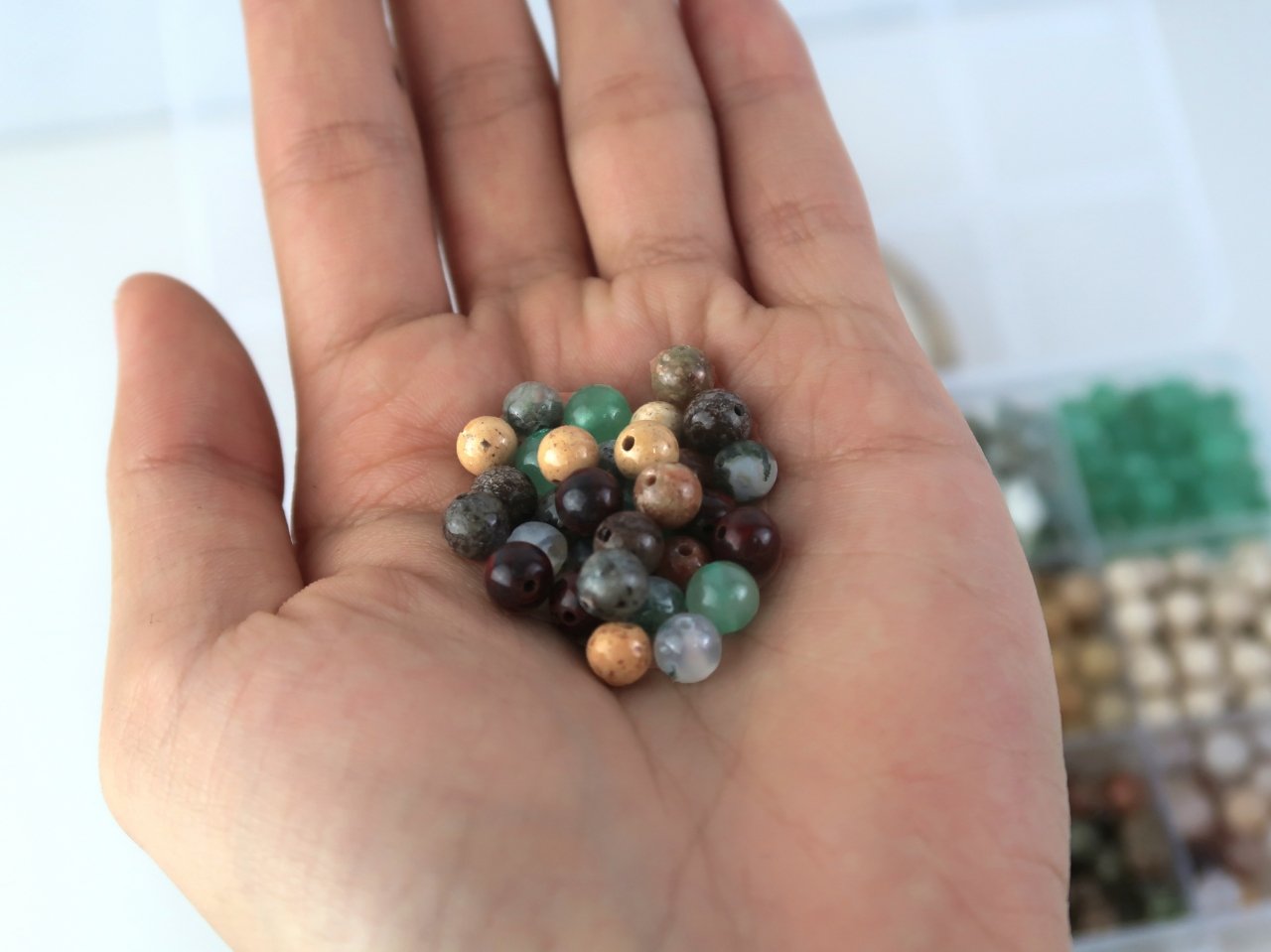 Hand holding a pile of assorted natural stone beads in various colors and patterns, with a blurred background of bead storage containers.
