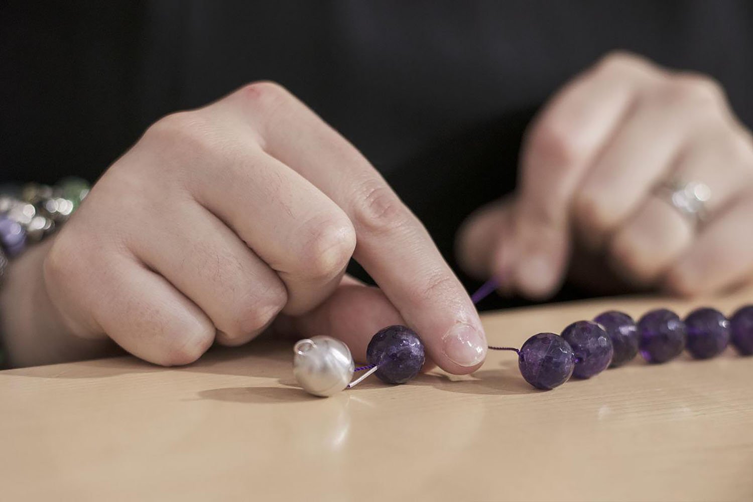 Hands stringing purple gemstone beads onto a purple thread, with a silver bead in the foreground.