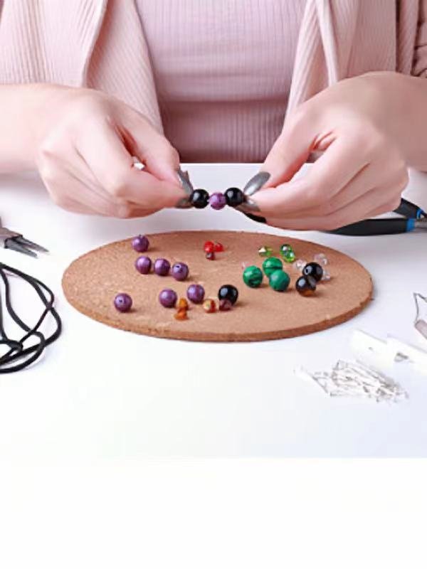 Hands stringing black and purple beads onto a thin cord, with various colorful beads scattered on a brown circular mat.