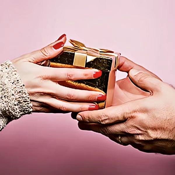 Two hands exchanging a small gift box tied with a gold ribbon against a pink background.