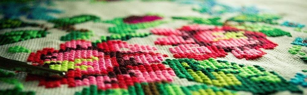 Close-up of cross-stitch embroidery with pink and green floral patterns, showing needle and thread detail.