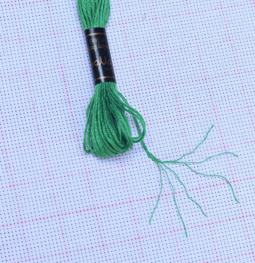 Green embroidery floss wound on a black spool with gold trim, resting on white cross-stitch fabric.