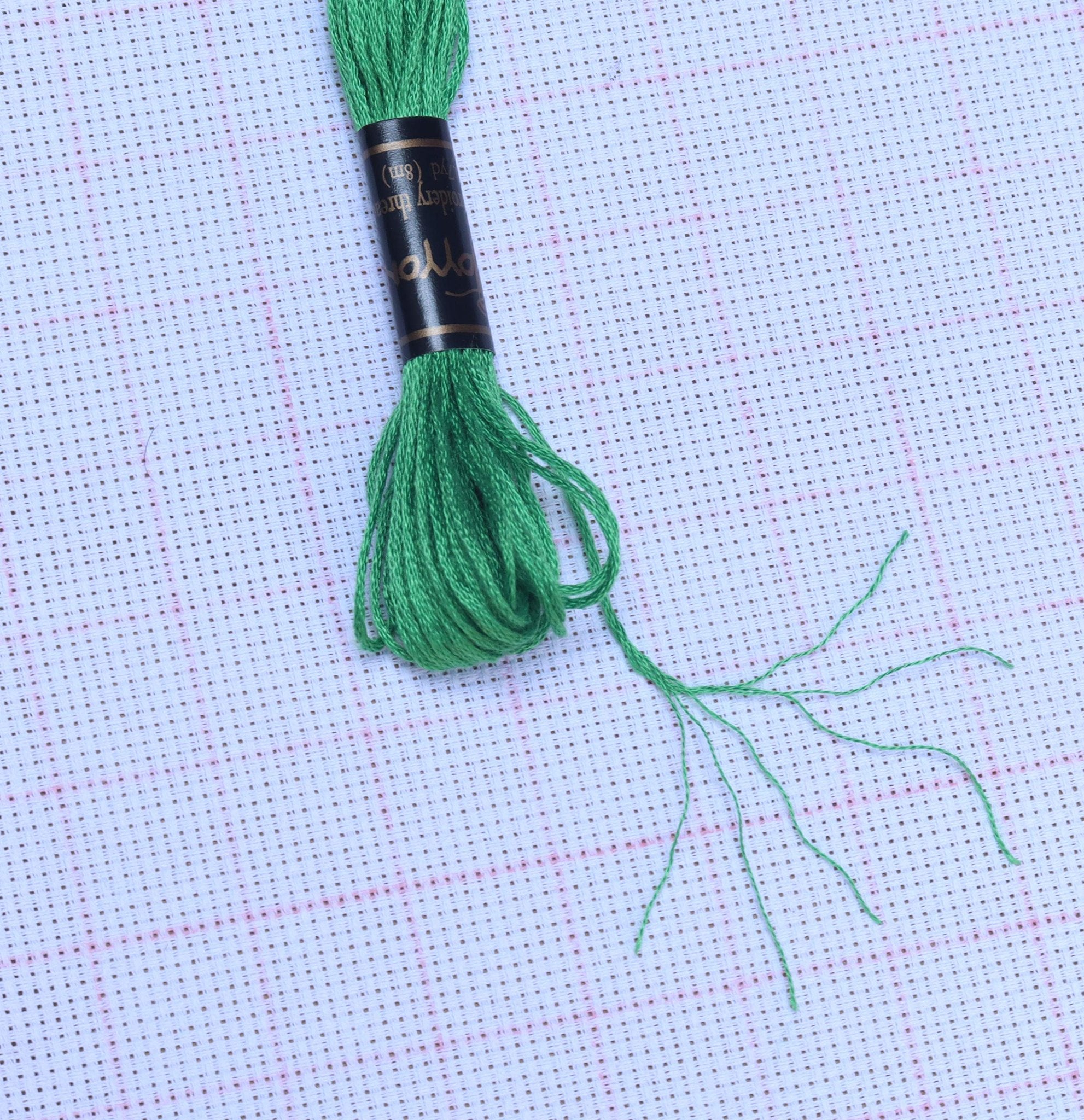 Green embroidery floss wound on a black spool with gold trim, resting on white cross-stitch fabric.
