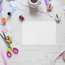 Craft supplies including scissors, thread, ribbon, and paper arranged on a light wooden surface.