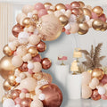 Blush pink and gold balloon garland arch with confetti balloons, set against a backdrop with decorative cakes and dried pampas grass.