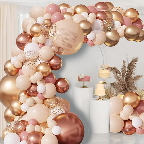 Blush pink and gold balloon garland arch with confetti balloons, set against a backdrop with decorative cakes and dried pampas grass.