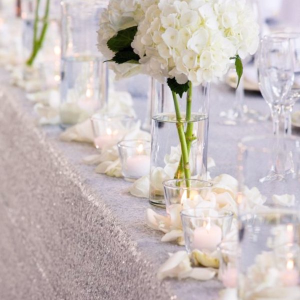 Table decorated with white hydrangea centerpieces, candles, and scattered white petals.