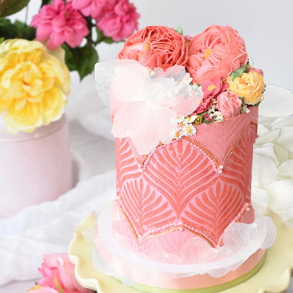 Decorated pink cake with floral frosting and edible lace accents, set against a backdrop of pink and yellow flowers.