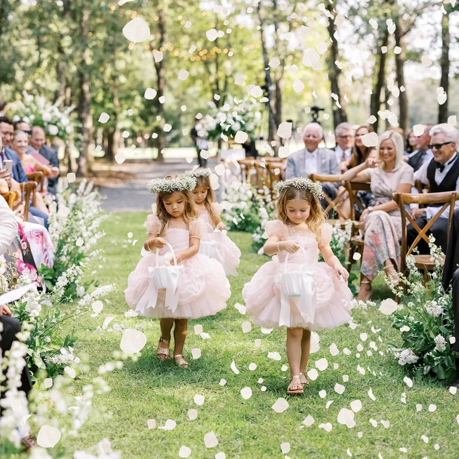 Flower girls in pink dresses walk down a grassy aisle scattering white petals at a wedding ceremony.