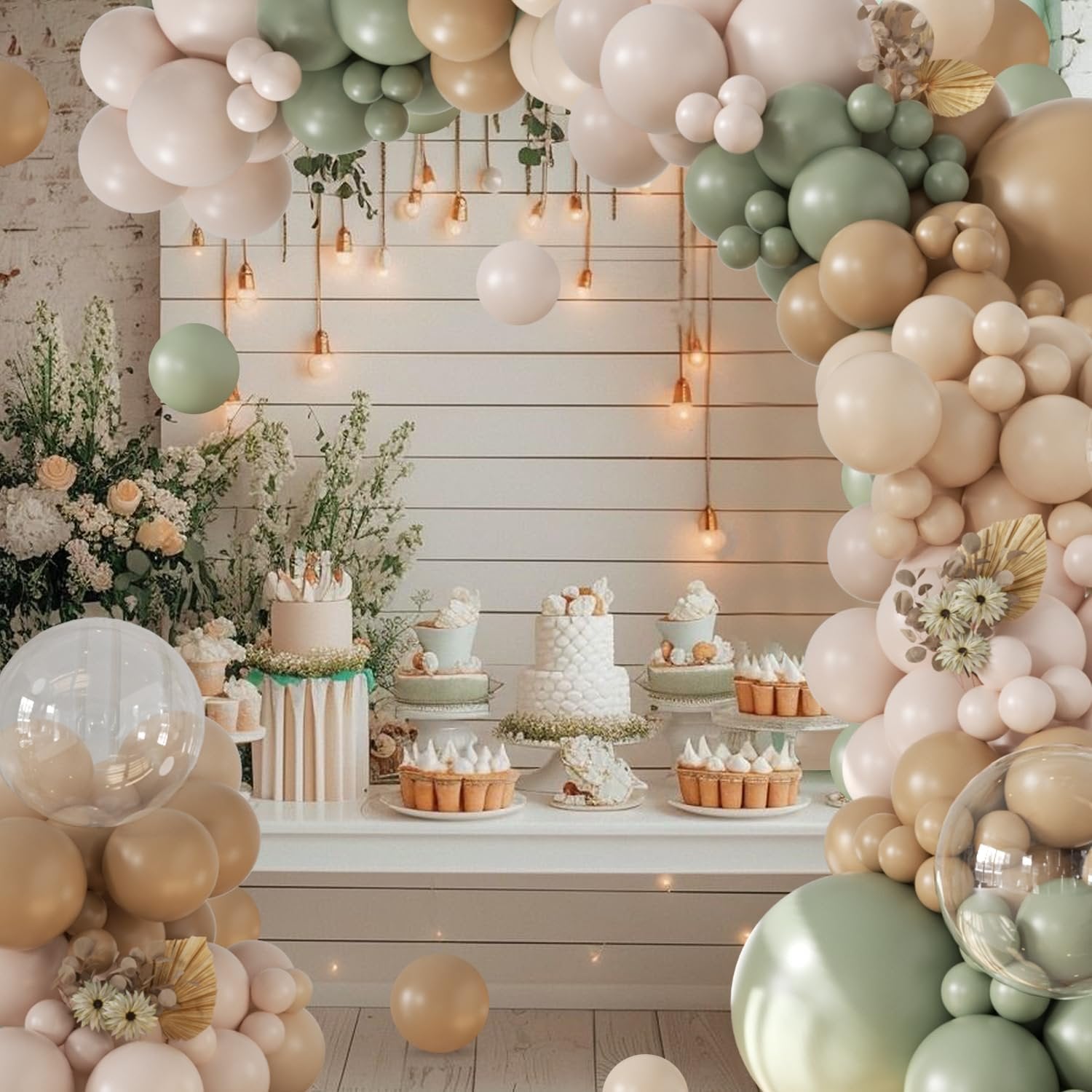 Sage green and neutral balloon arch over a dessert table with cakes and tarts, decorated with flowers and string lights.