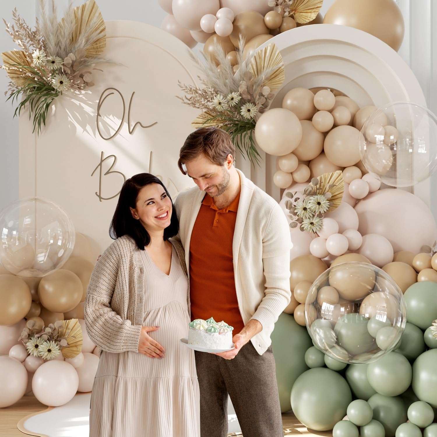 Pregnant couple holding a cake in front of a balloon arch with 'Oh Baby' text