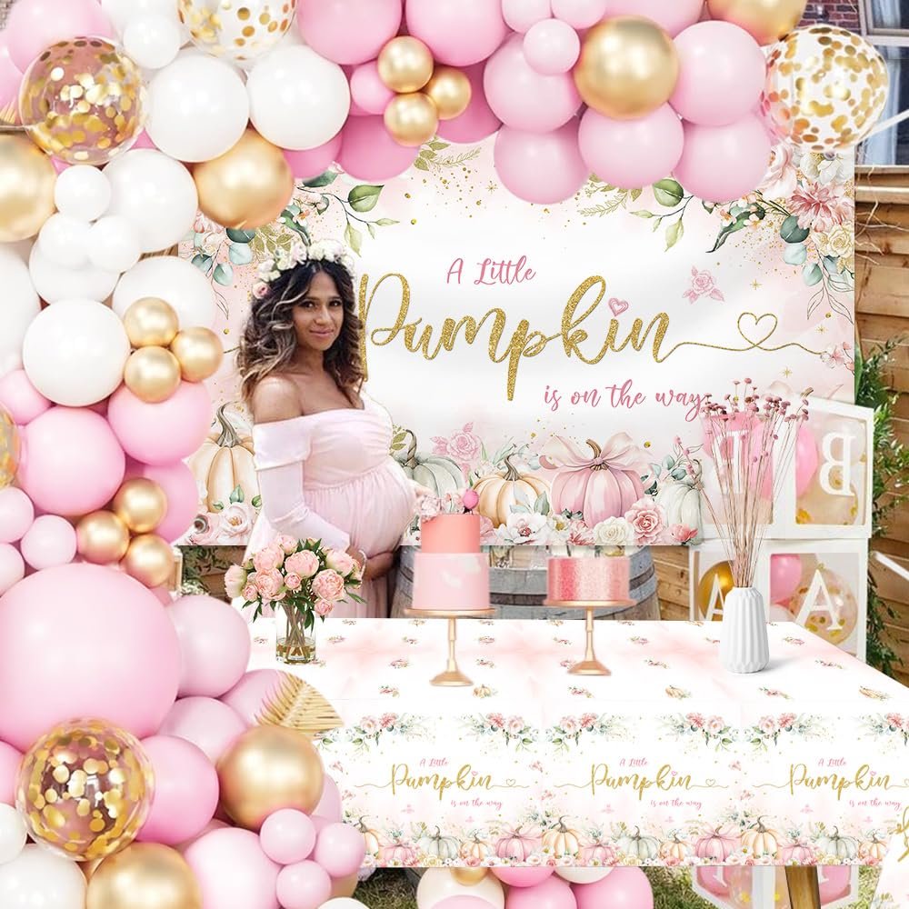 Pregnant woman posing at a baby shower with pink and gold balloons, a floral banner, pumpkins, and cakes.