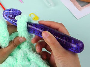 Hand holding a large purple glitter crochet hook with a light green fuzzy yarn.