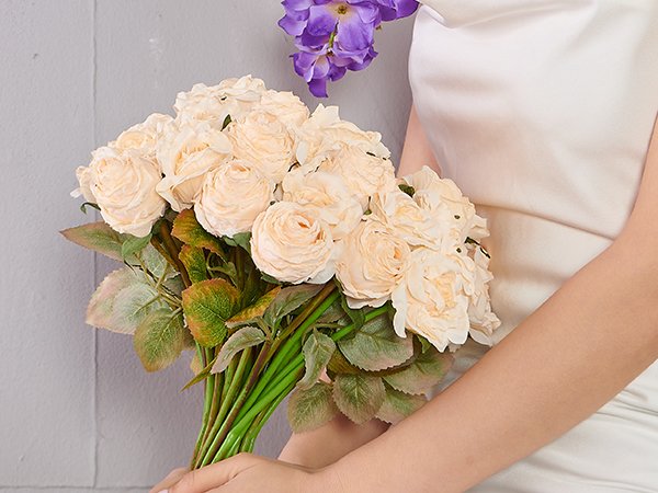 Bunch of cream artificial roses with green stems and leaves held by hands, with purple flowers in background.