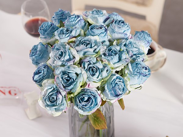 Bouquet of blue artificial roses in a glass vase on a white tablecloth, with a wine glass in the background.