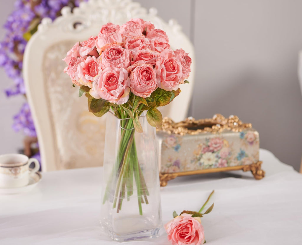 Bouquet of pink artificial roses in a clear vase on a table with a decorative box and teacup.