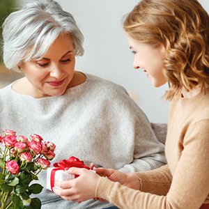 Young woman gives a gift to an older woman, with a bouquet of pink roses nearby.