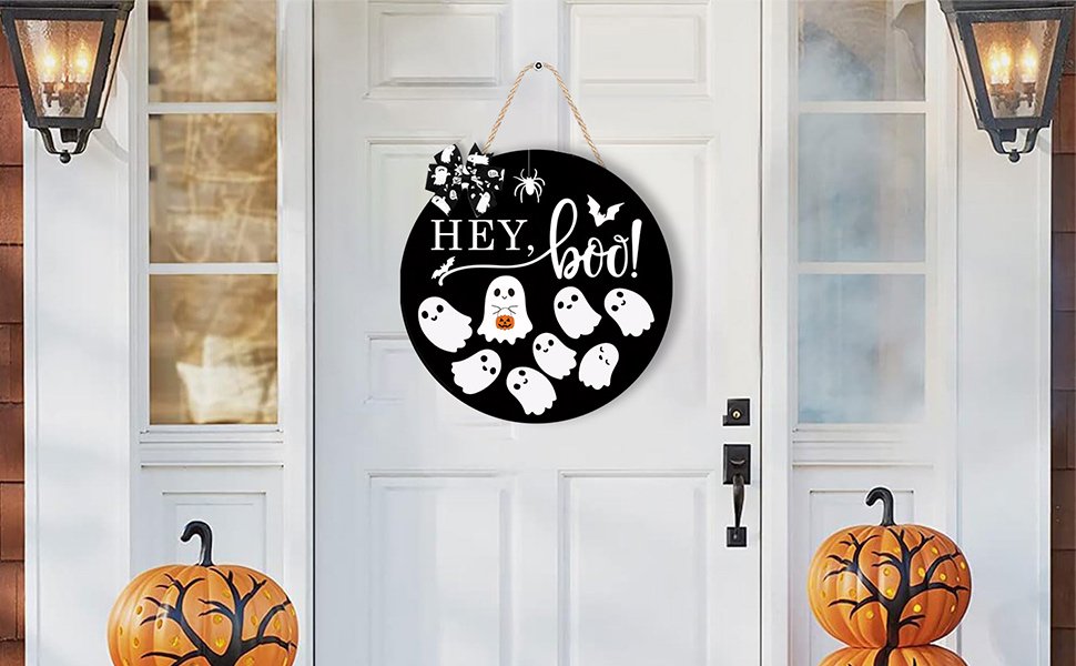 Round black Halloween door sign with 'Hey, Boo!' text and ghost graphics, hanging on a white door between two lanterns and pumpkins.