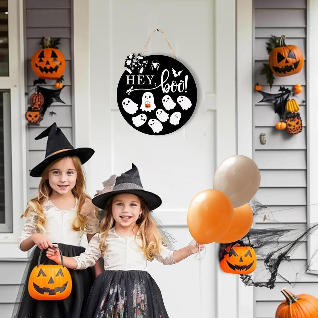 Two young girls in witch costumes stand on a porch decorated for Halloween, holding candy buckets and balloons. A round wooden sign with ghosts and the words 'Hey Boo' hangs on the door.