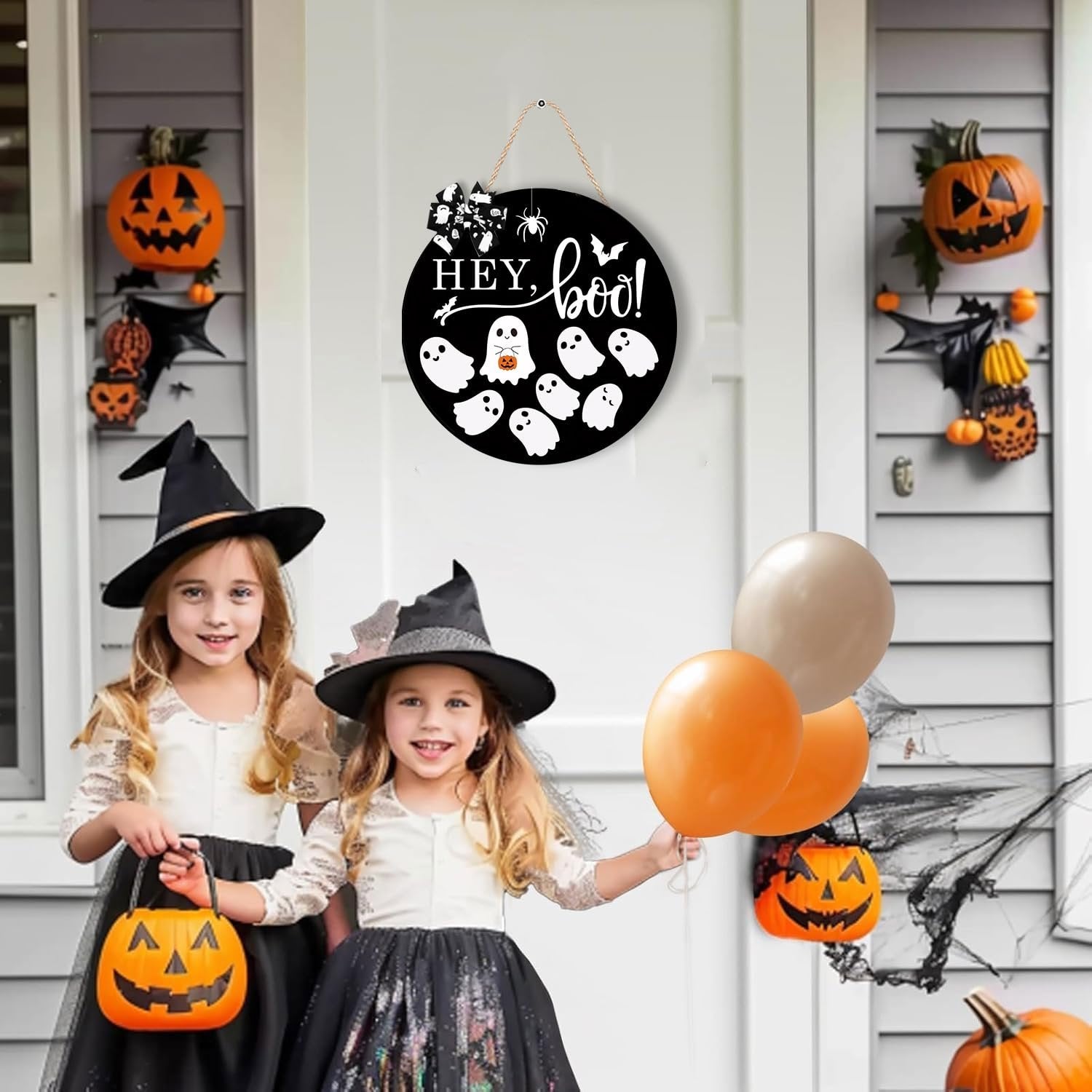 Two young girls in witch costumes stand on a porch decorated for Halloween, holding candy buckets and balloons. A round wooden sign with ghosts and the words 'Hey Boo' hangs on the door.
