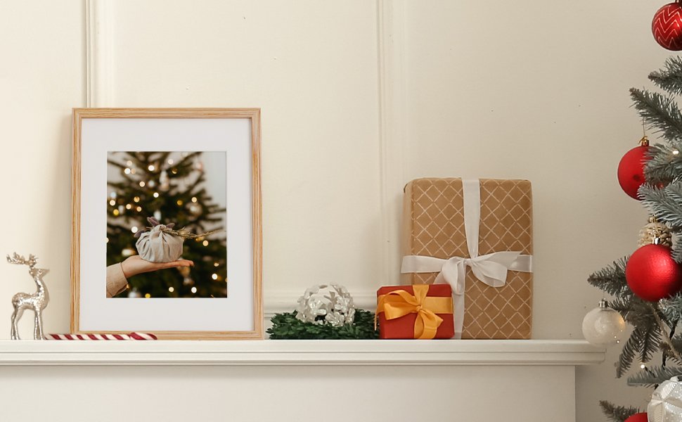 Wooden picture frame displaying a hand holding a fabric-wrapped gift in front of a blurred Christmas tree, on a shelf with gifts and ornaments.
