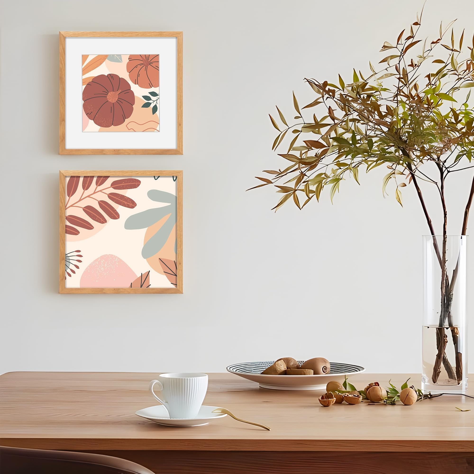 Two framed abstract art prints with pumpkins and leaves hanging on a wall above a wooden table with a coffee cup and vase.