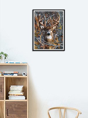 Framed 5D diamond art painting of a deer with large antlers peeking through camouflage foliage, displayed on a wall above a bookshelf.