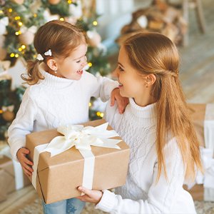 Mother and daughter in white sweaters holding a wrapped gift box with a white ribbon in front of a decorated Christmas tree.