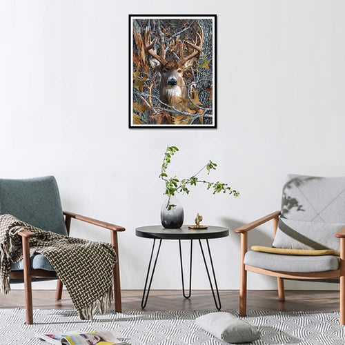 Diamond art painting of a deer with large antlers camouflaged in a forest setting, framed and hanging on a wall above a small table.