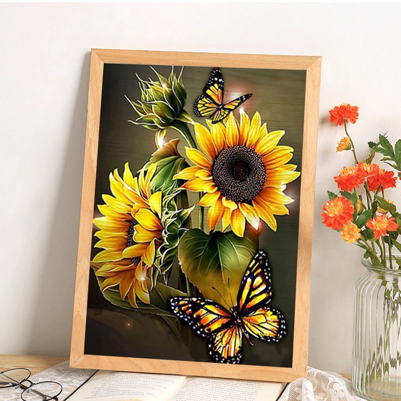 Diamond painting kit featuring sunflowers and butterflies in a wooden frame, displayed on a book with glasses nearby.
