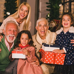 Multi-generational family smiling and holding Christmas gifts, with festive decorations in the background.
