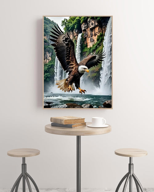 Framed diamond painting of a bald eagle in flight over waterfalls, displayed on a wall above a small table with books and a coffee cup.