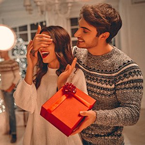 Man surprising woman with a red gift box, both smiling in a festive indoor setting with Christmas lights.