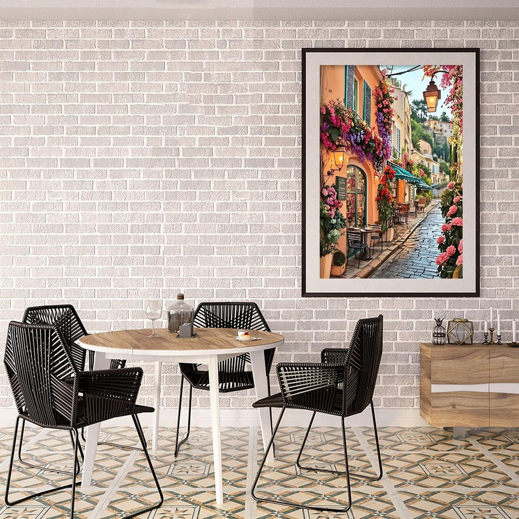 Framed diamond painting of a flower-lined street in a European town, displayed above a dining table with chairs.