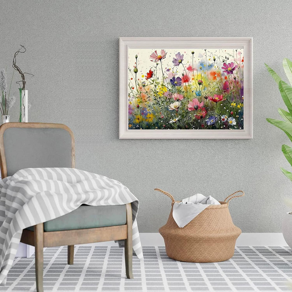 Framed watercolor painting of a field of colorful wildflowers hanging on a textured gray wall above a chair and basket.