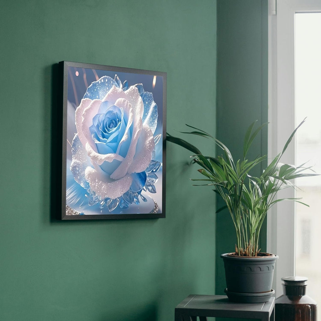 Framed diamond painting of a blue and white rose with sparkling details, hanging on a green wall next to a potted plant.