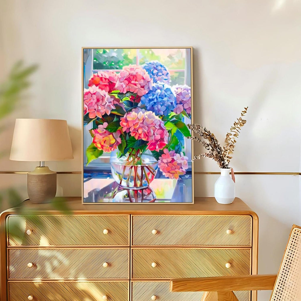 Framed diamond painting of pink and blue hydrangeas in a glass vase on a dresser, next to a lamp and dried flowers.