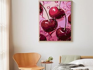 Framed diamond painting of glossy red cherries with pink water droplets hanging on a wall above a wooden chair and bed.