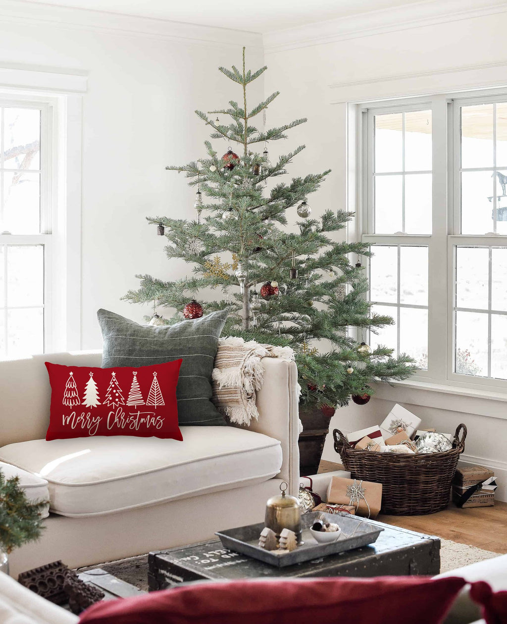 Red Christmas pillow with white trees and 'Merry Christmas' text on a couch next to a decorated tree.