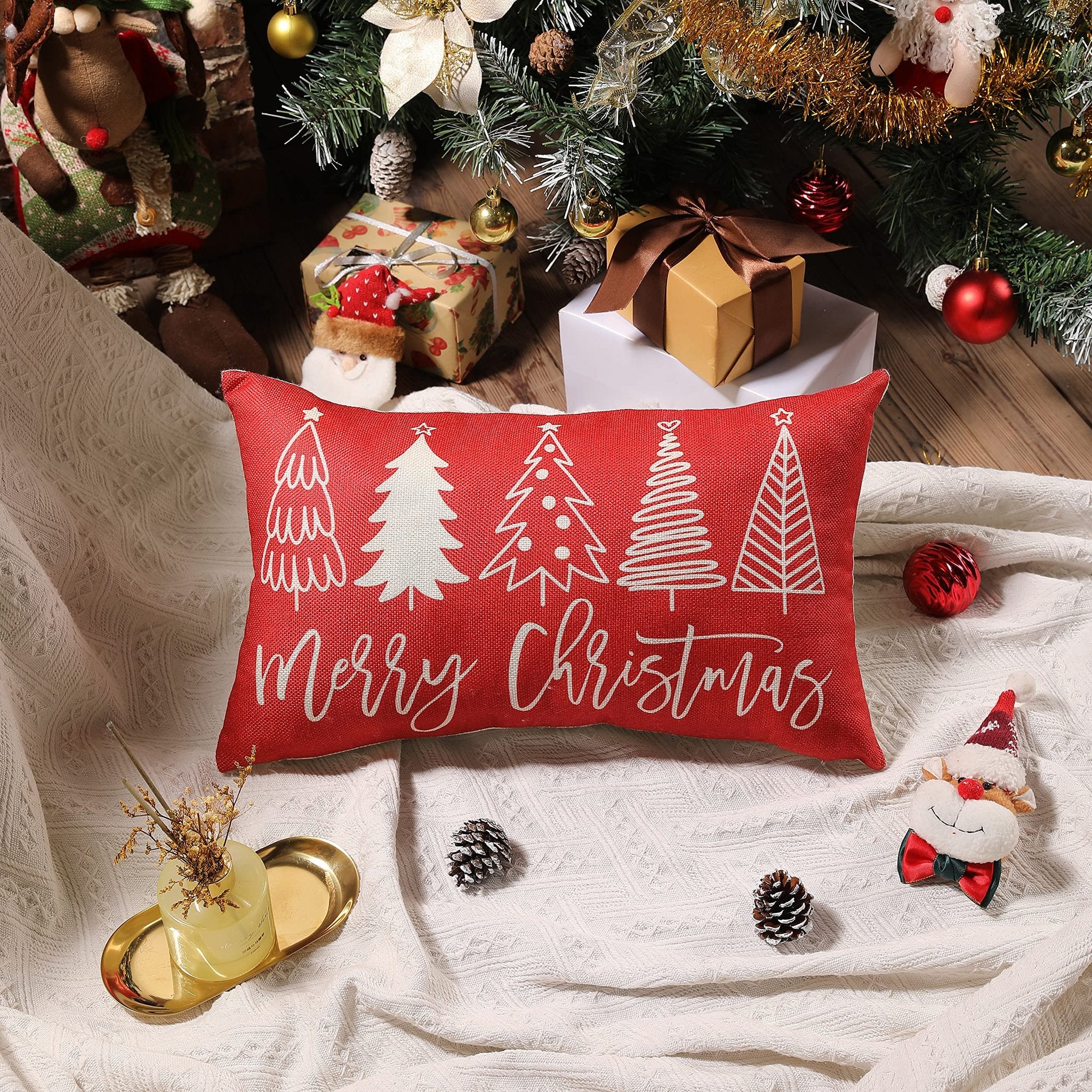 Red lumbar pillow with white Christmas trees and 'Merry Christmas' text, set on a white blanket under a decorated Christmas tree.