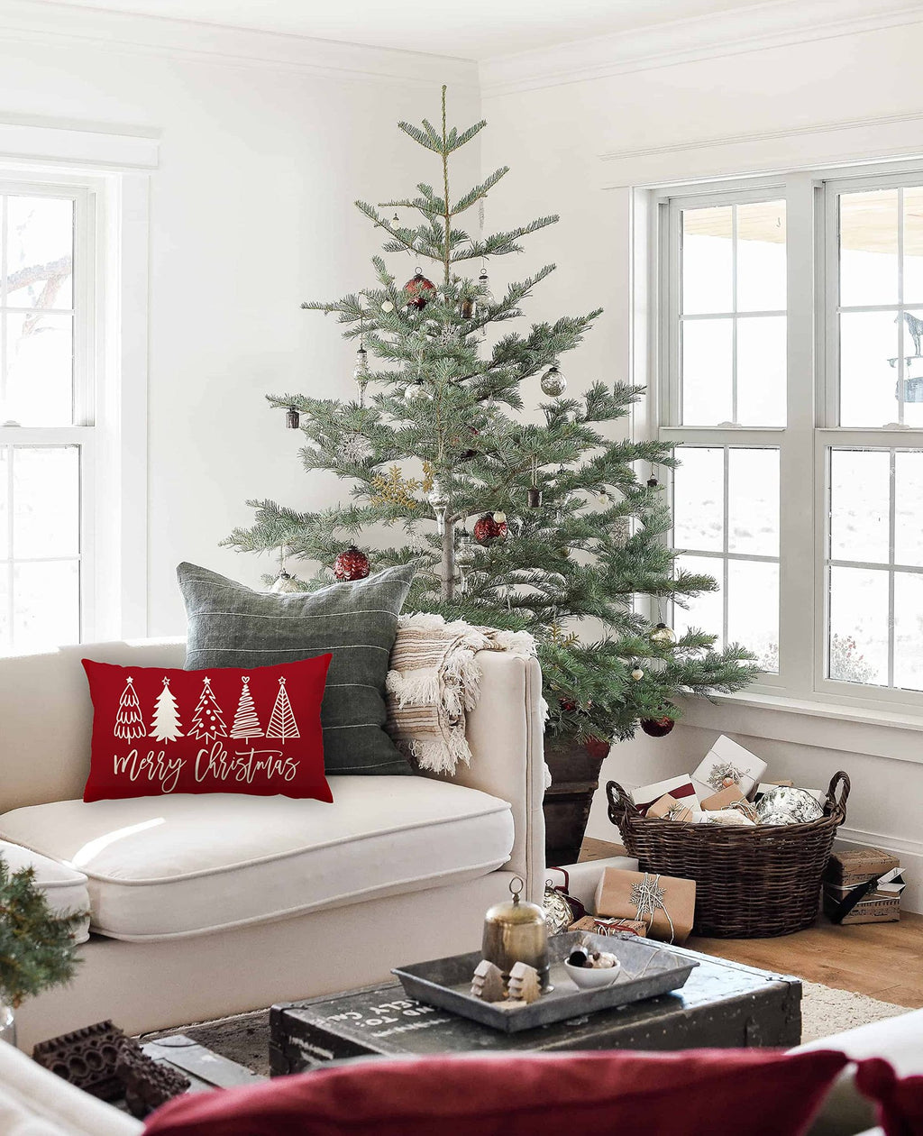 Red lumbar pillow with white Christmas trees and 'Merry Christmas' text on a couch next to a Christmas tree.